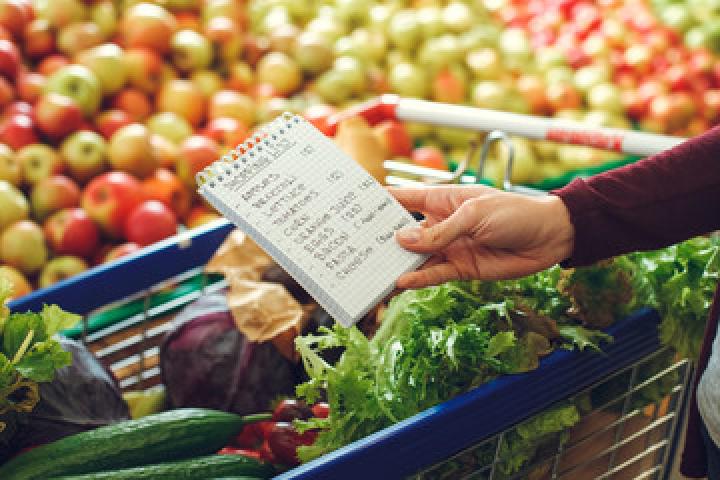 Photo of a hand holding a shopping list, above a shopping cart that is in front a section of yellow and red tomatoes